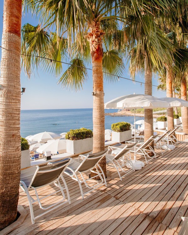 A wooden deck with lounge chairs and umbrellas, overlooking a beach with palm trees and a clear blue sky.