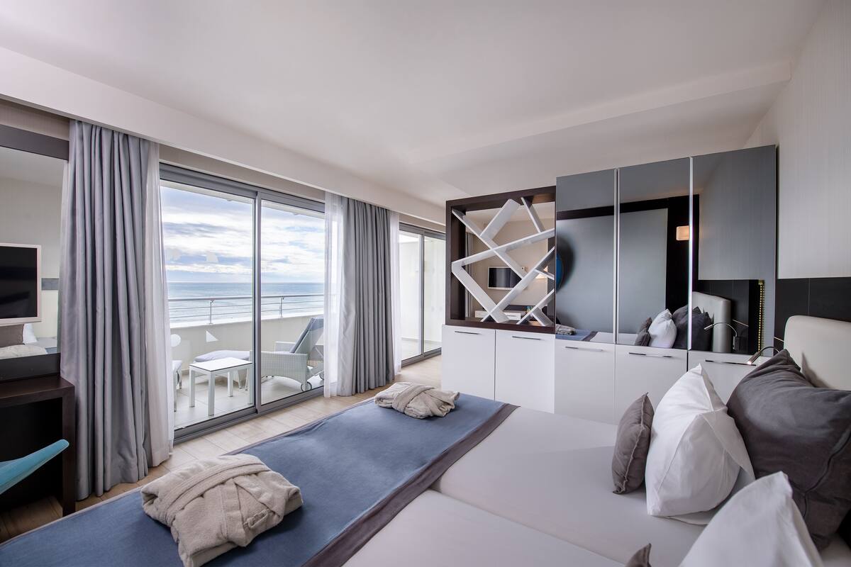 A modern hotel room with a large balcony, a bed with multiple pillows, a TV, and a seating area with a table and chairs.