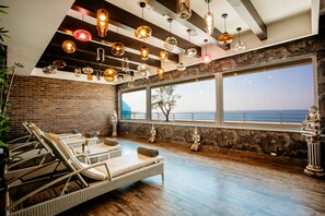 Ett spa-rum med flätade loungestolar, ett stort fönster med utsikt över havet och olika typer av hängande taklampor.