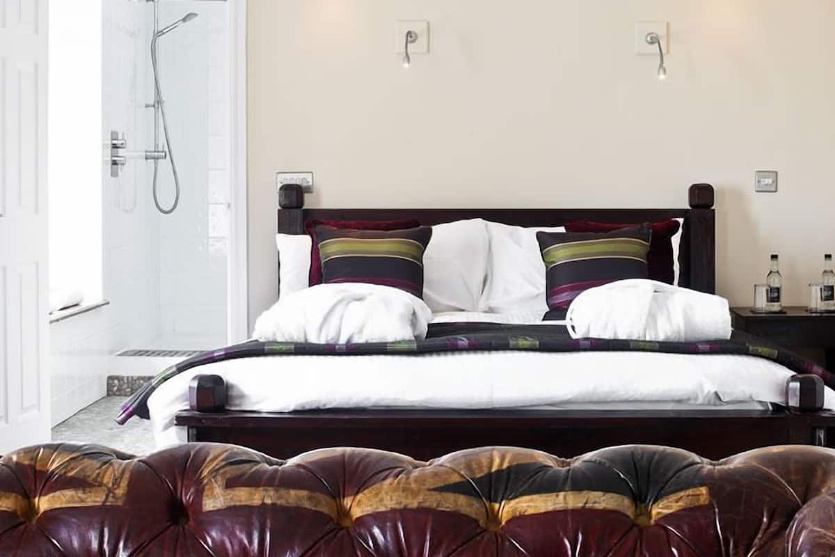 Cama bem arrumada com manta e travesseiros listrados, cabeceira de madeira, mesa de cabeceira com uma garrafa e um copo, e área de chuveiro com porta de vidro.
