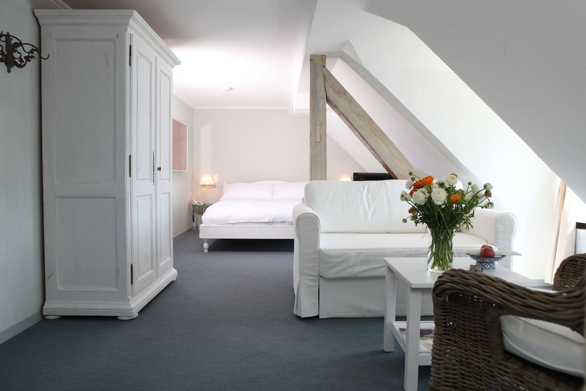 A bedroom with a bed, a white wardrobe, a white sofa, a wicker chair, and a small table with a vase of flowers.