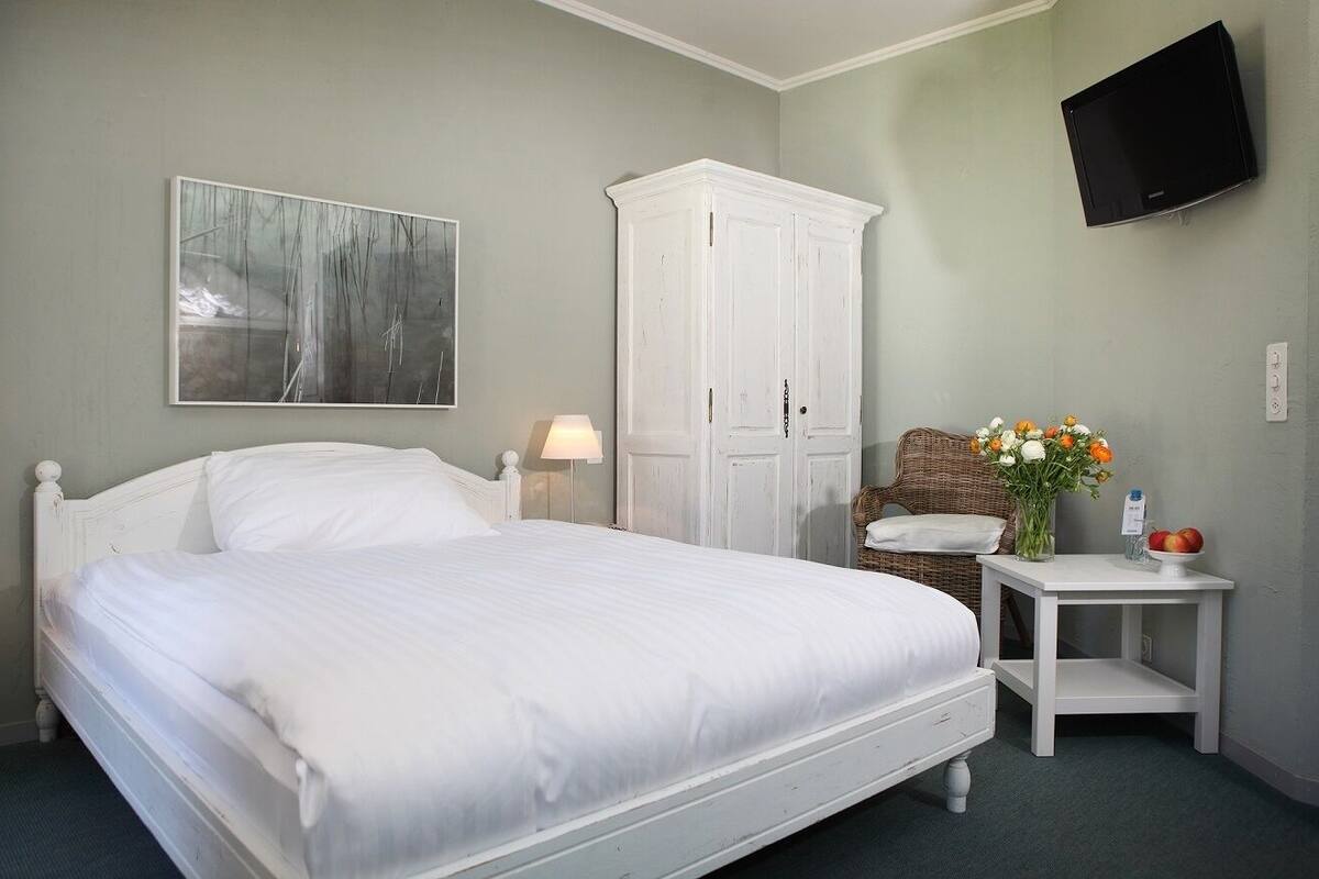 A neatly made bed in a room with a white wardrobe, a flat-screen TV mounted on the wall, and a small table with a vase of flowers.