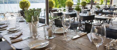 Une table à manger dressée pour un repas, avec des assiettes, des couverts et des verres, entourée de chaises et de plantes en pot, et en arrière-plan, une vue sur une étendue d’eau.