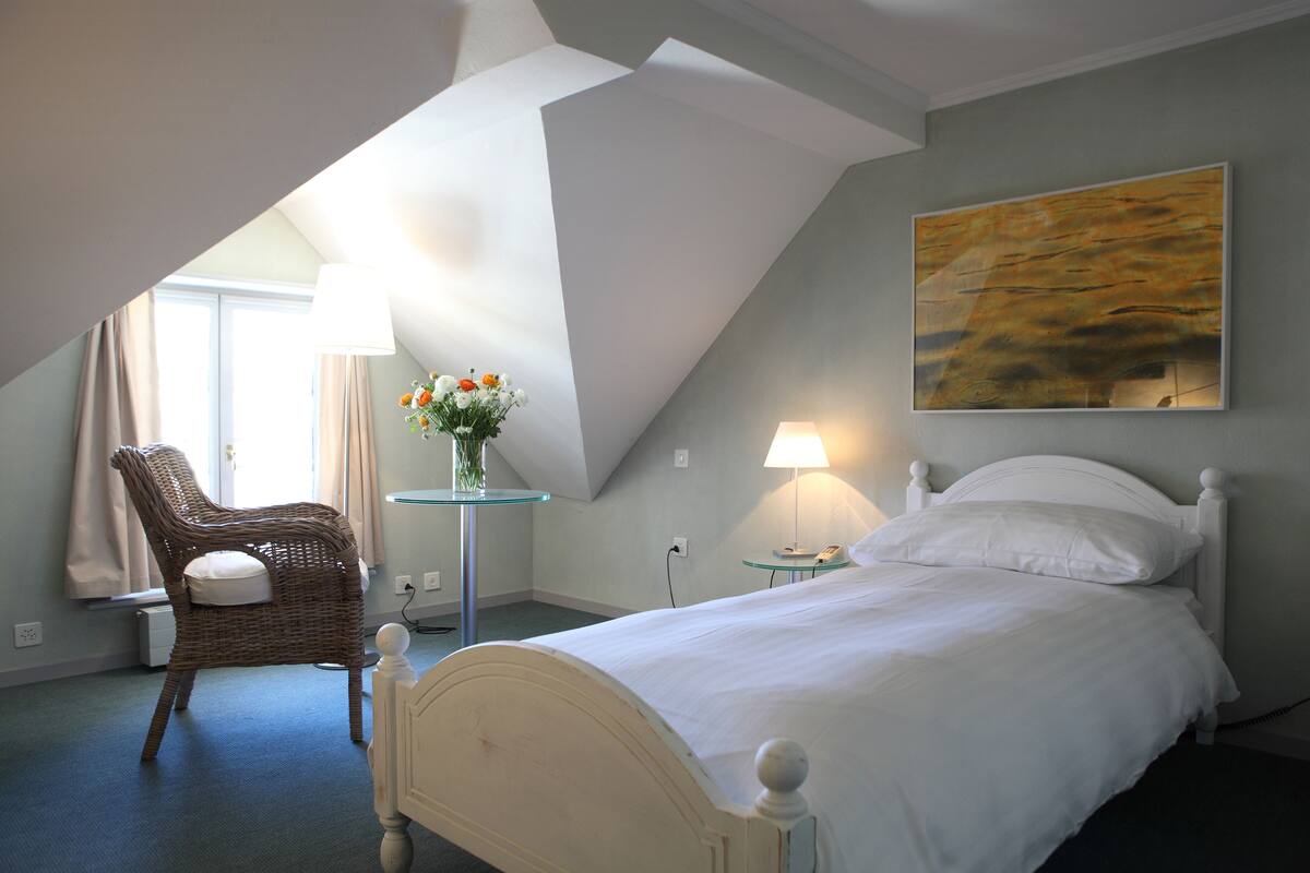 A bedroom with a bed, a chair, a table with a lamp, and a painting on the wall.