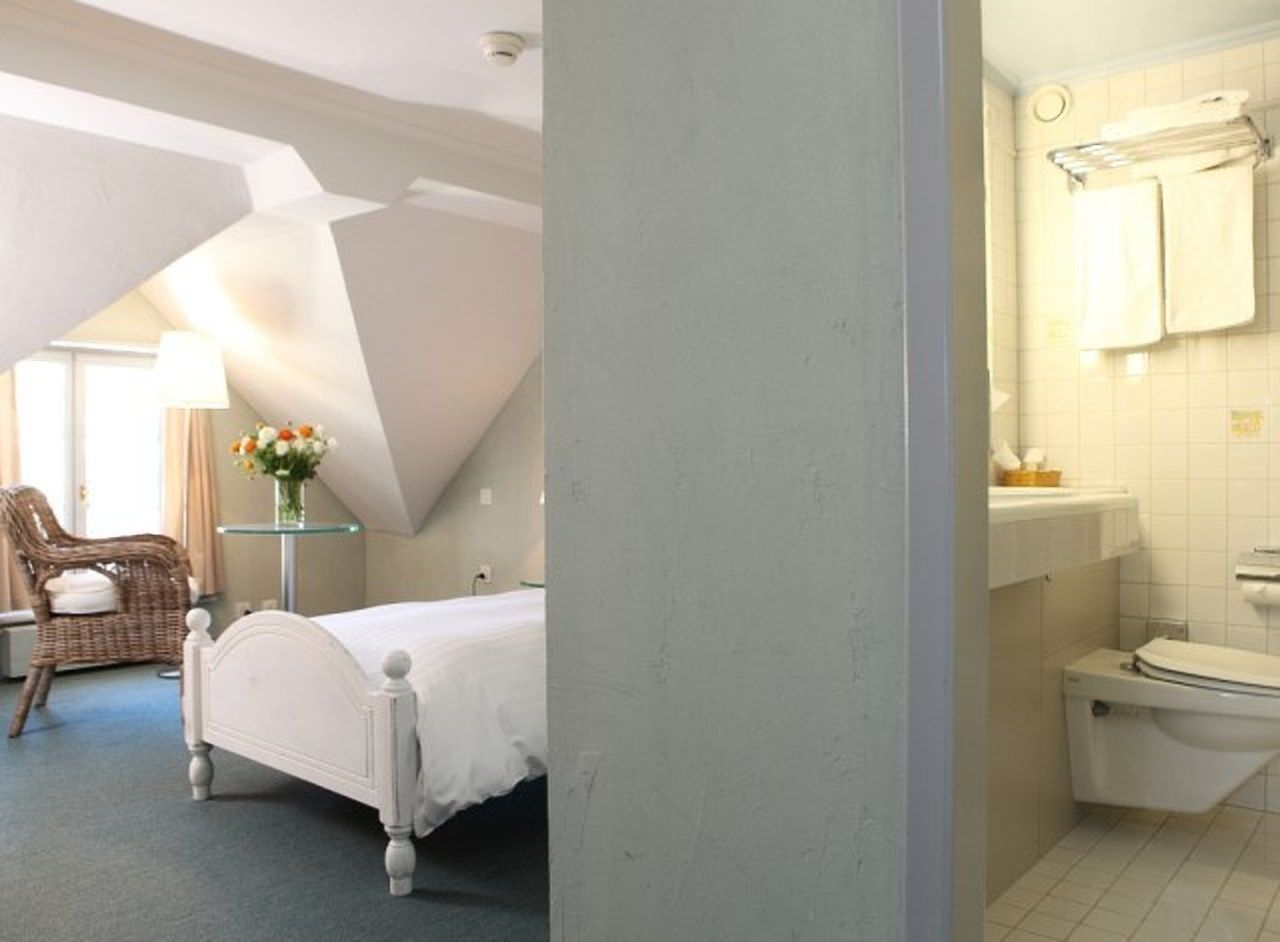 A bedroom with a bed, a chair, a small table with a vase of flowers, and a bathroom with a toilet and sink.