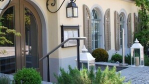 Entrada de un edificio con puerta de cristal, escalera metálica y un pequeño jardín con plantas y una silla blanca.
