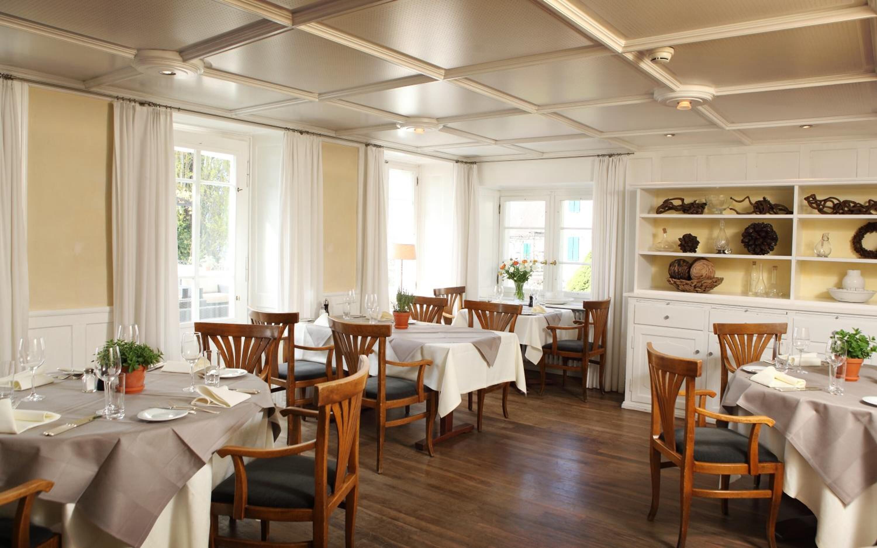A dining area with multiple tables set for guests, wooden chairs, and a white decorative shelf with various items.