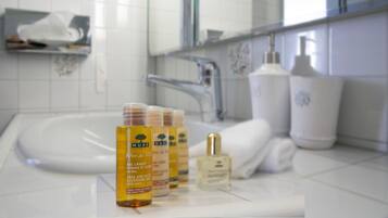 A bathroom with a sink, a mirror, and toiletries including a bottle of Nuxe body lotion and a bottle of perfume.