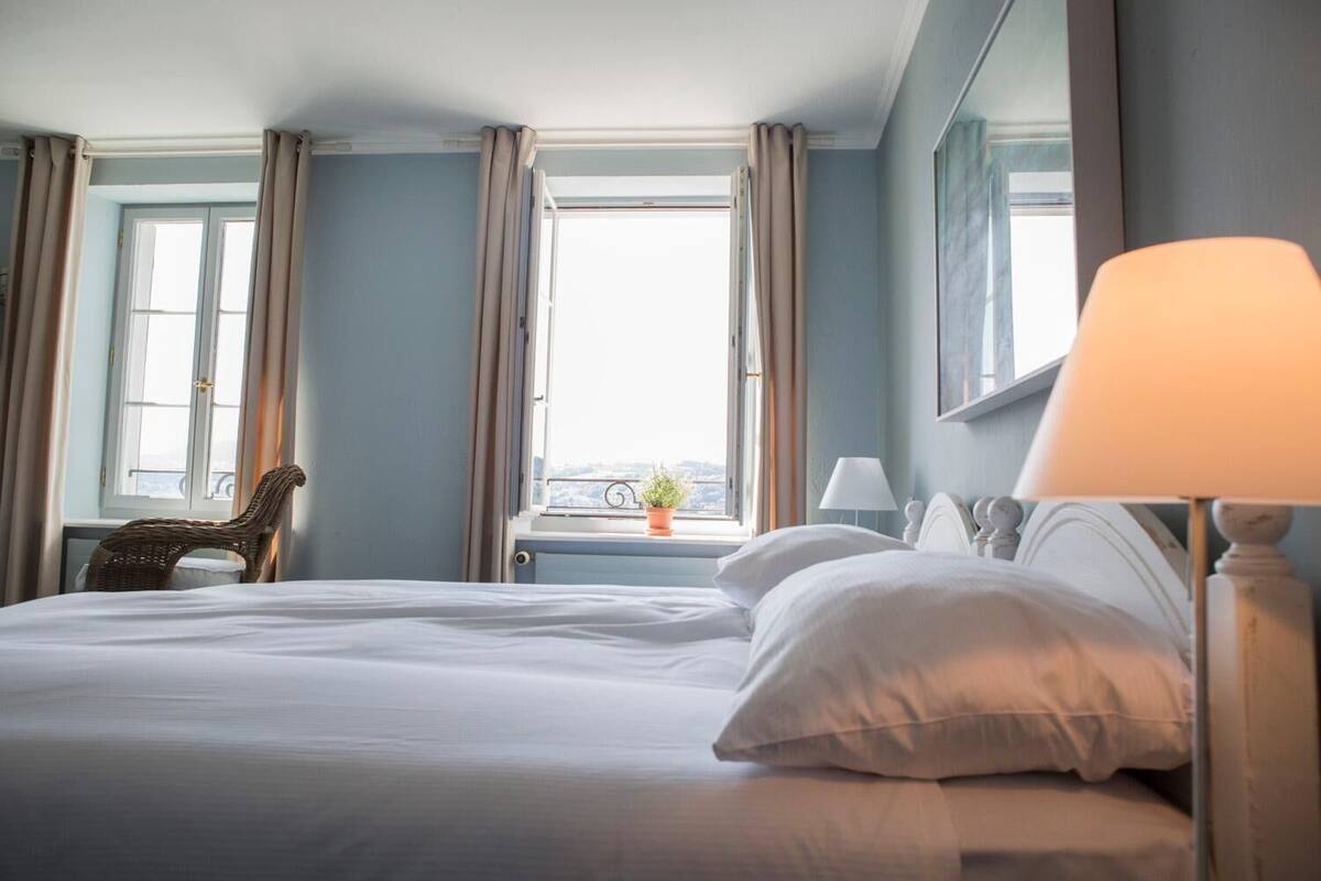 A hotel room with a large bed, a chair, a window with curtains, and a lamp.