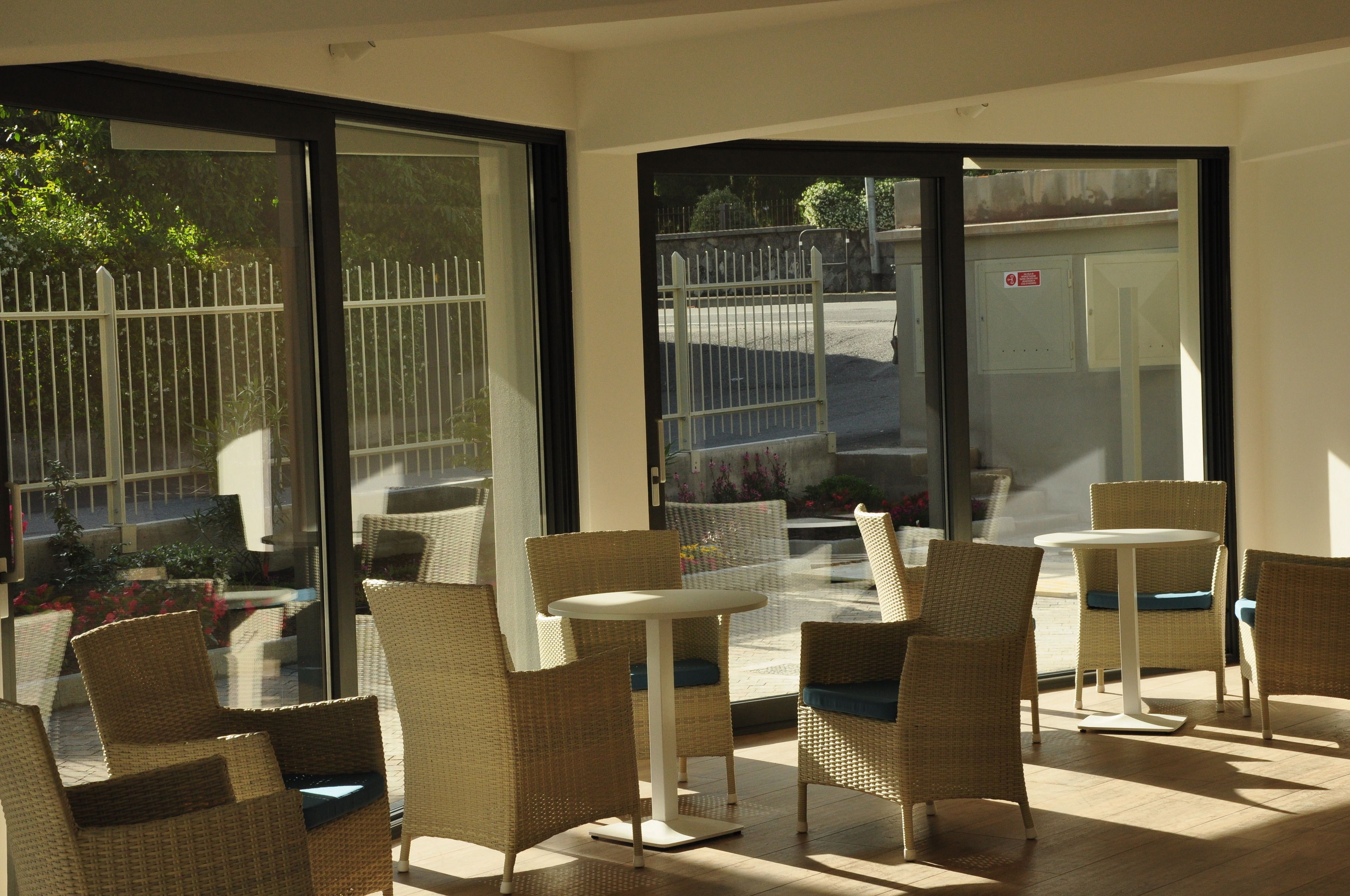 A sunlit indoor seating area with wicker chairs and a small round table, large glass doors leading outside, and a view of a fenced garden.