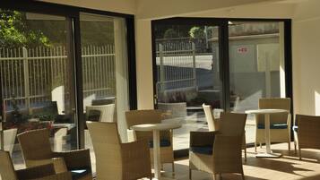 A sunlit indoor seating area with wicker chairs and a small round table, large glass doors leading outside, and a view of a fenced garden.