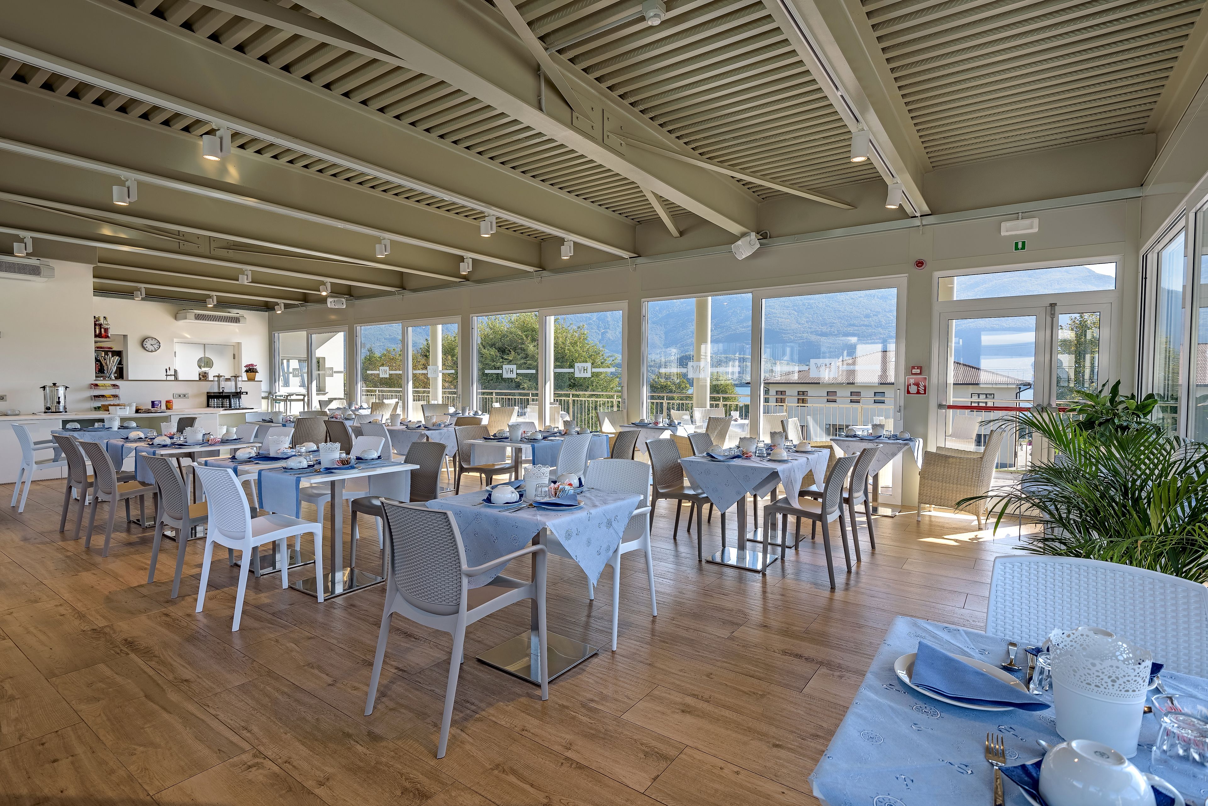 A spacious dining area with multiple tables and chairs, large windows offering views of the outside, and a modern ceiling design.