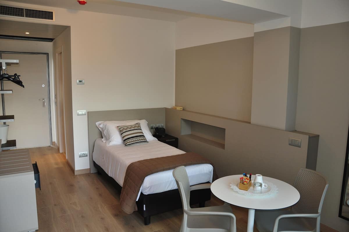 Une chambre d’hôtel de taille réduite, comprenant un lit simple, une petite table avec une chaise et un coin cuisine.