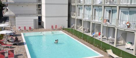 A hotel with a swimming pool surrounded by sun loungers and a covered area.