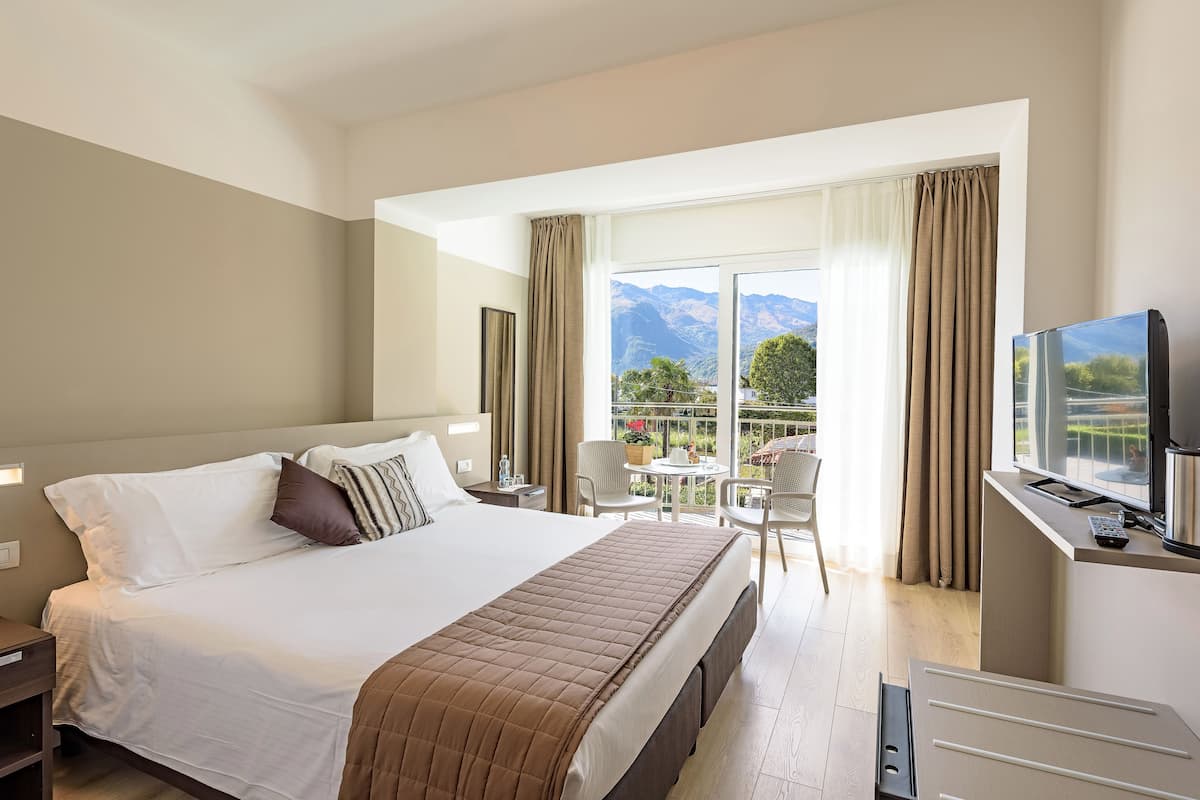 Une chambre d’hôtel avec un grand lit, un bureau avec une télévision, un balcon avec une table et des chaises, et une vue sur les montagnes.
