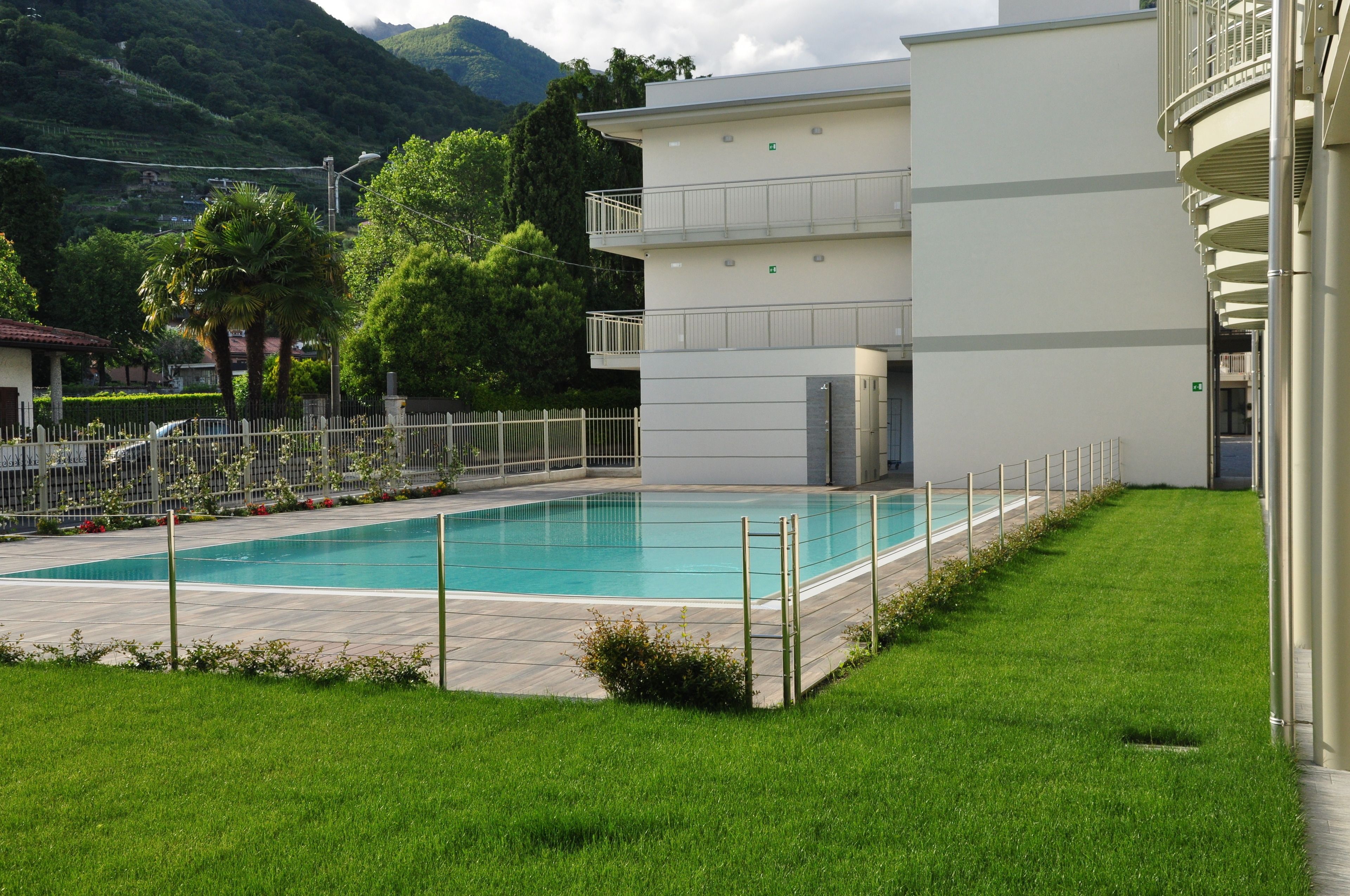 A swimming pool with a white fence, a grassy area, and a building with balconies.