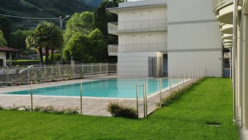 A swimming pool with a white fence, a grassy area, and a building with balconies.