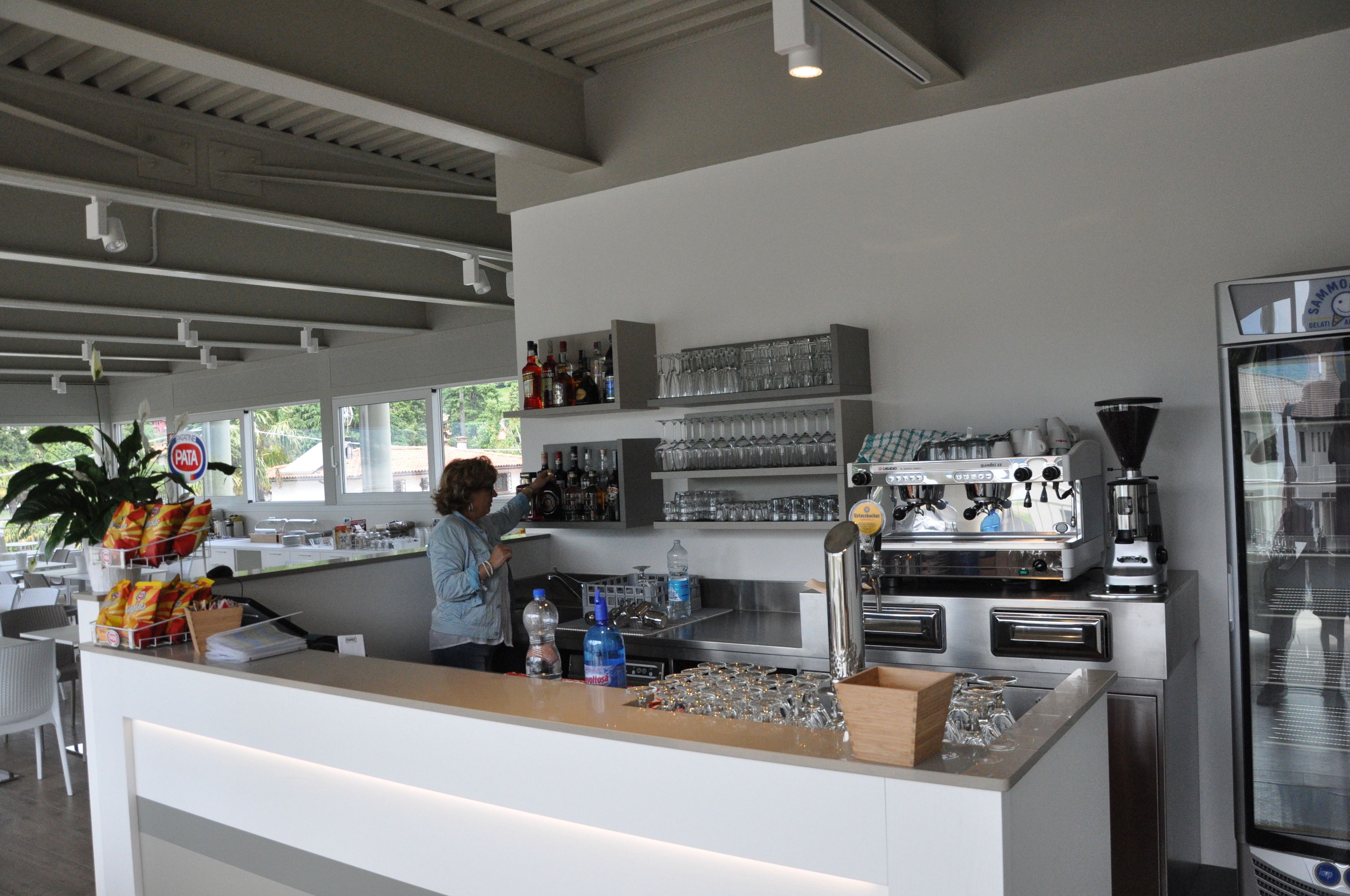 Zona de bar con mostrador, estanterías con botellas y vasos, una máquina de café y un refrigerador.