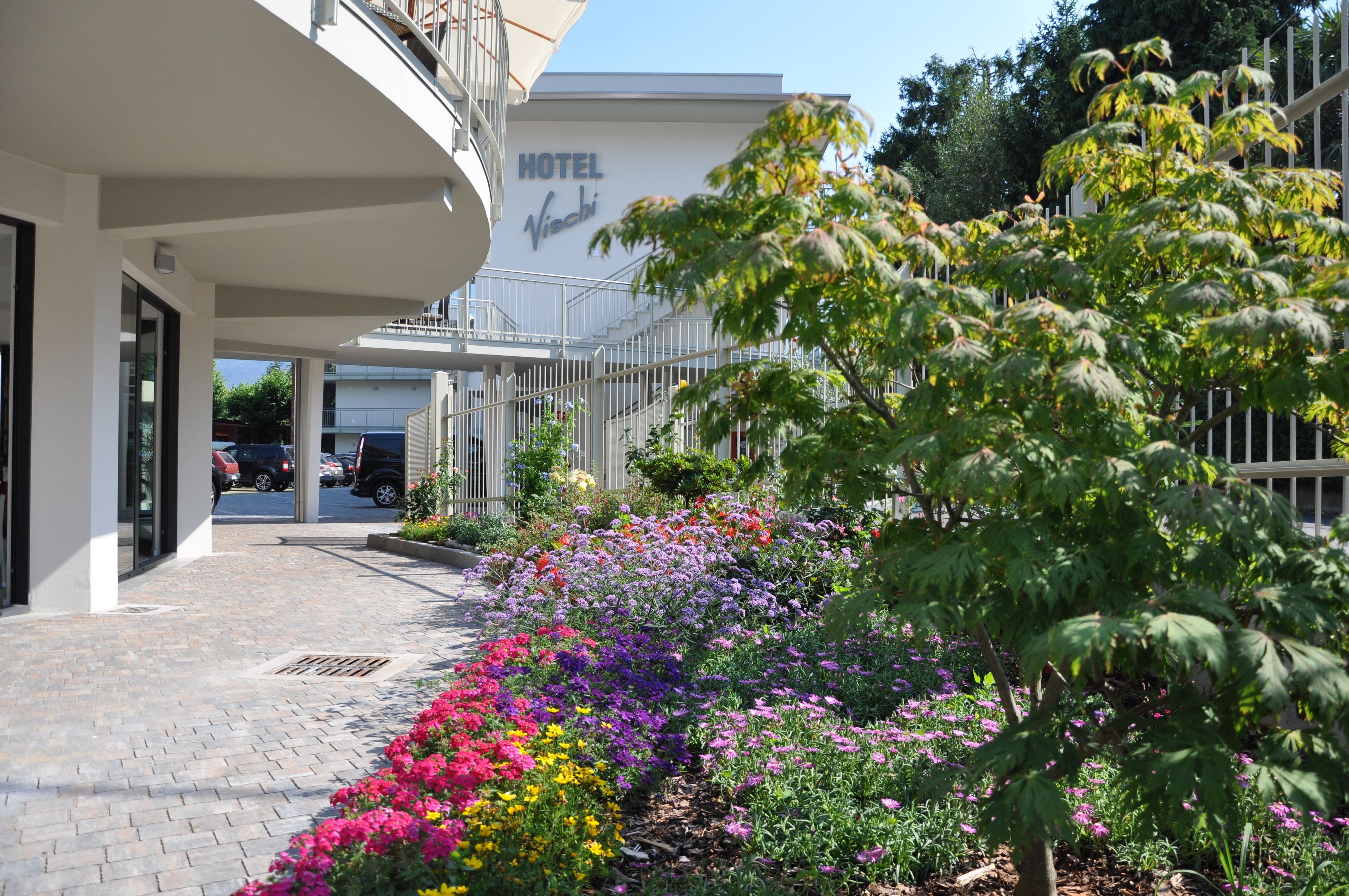 A hotel entrance with a paved walkway, a sign reading "HOTEL Visachi," and a variety of colorful flowers along the path.