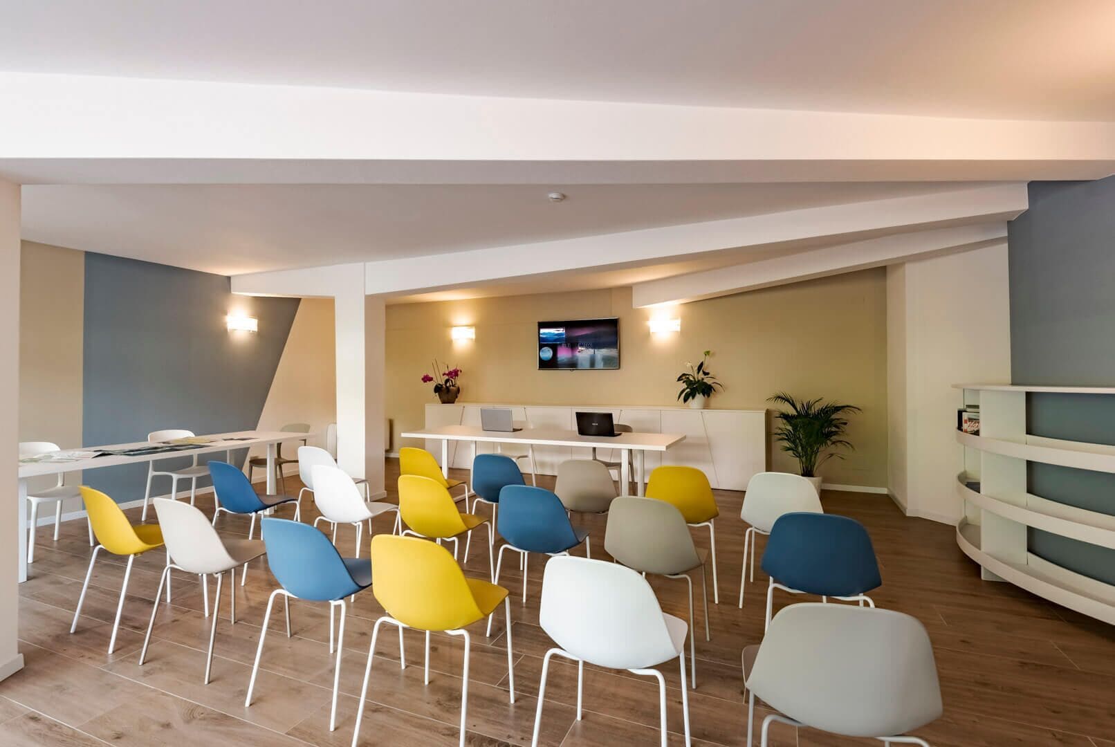 A modern meeting room with a long table, colorful chairs, a flat-screen TV, and a potted plant.