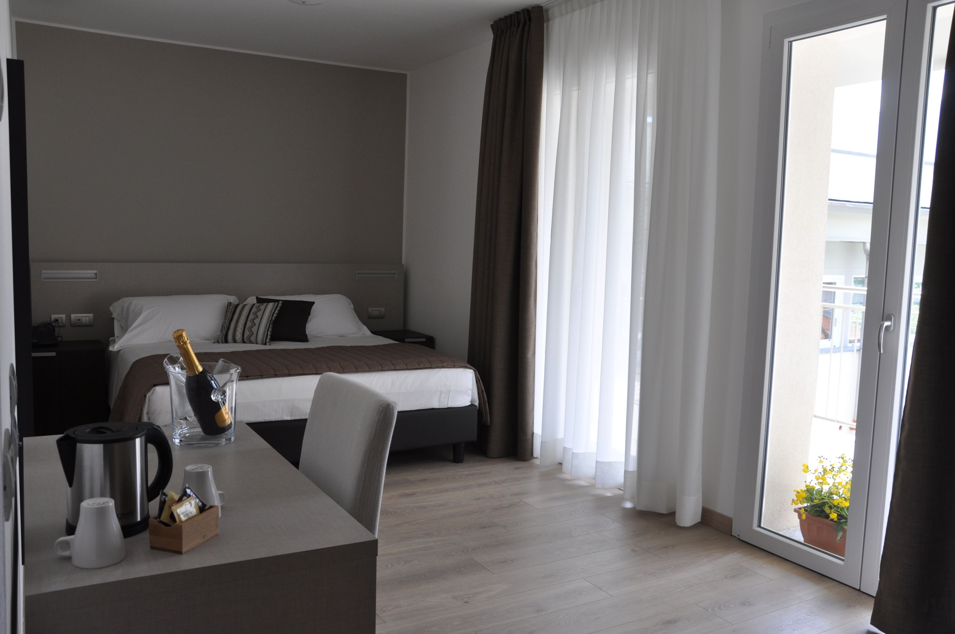 Habitación de hotel con cama, una mesa con cafetera, una botella de champán y una planta en maceta junto a la ventana.