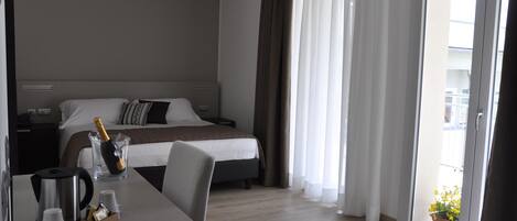 A hotel room with a bed, a table with a coffee maker, a bottle of champagne, and a potted plant by the window.