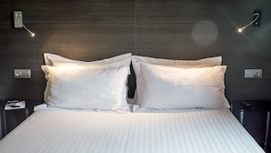 Una cama bien hecha con sábanas blancas y dos almohadas, un cabecero de madera oscura y dos lámparas de lectura fijadas a la pared.