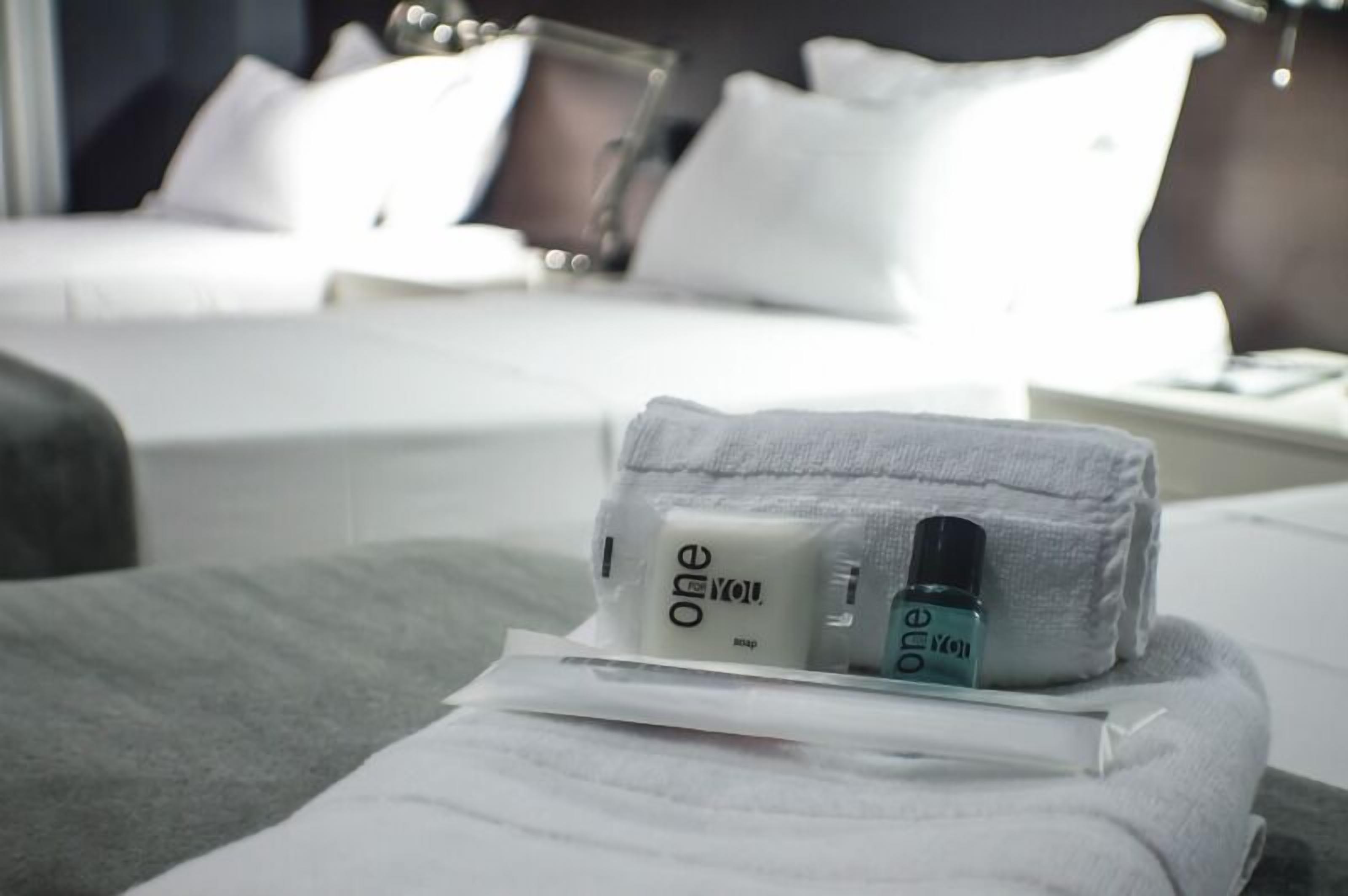 A neatly made bed with a white comforter and pillows, a small toiletry bag with a bottle and a bar of soap, and a white nightstand with a lamp.