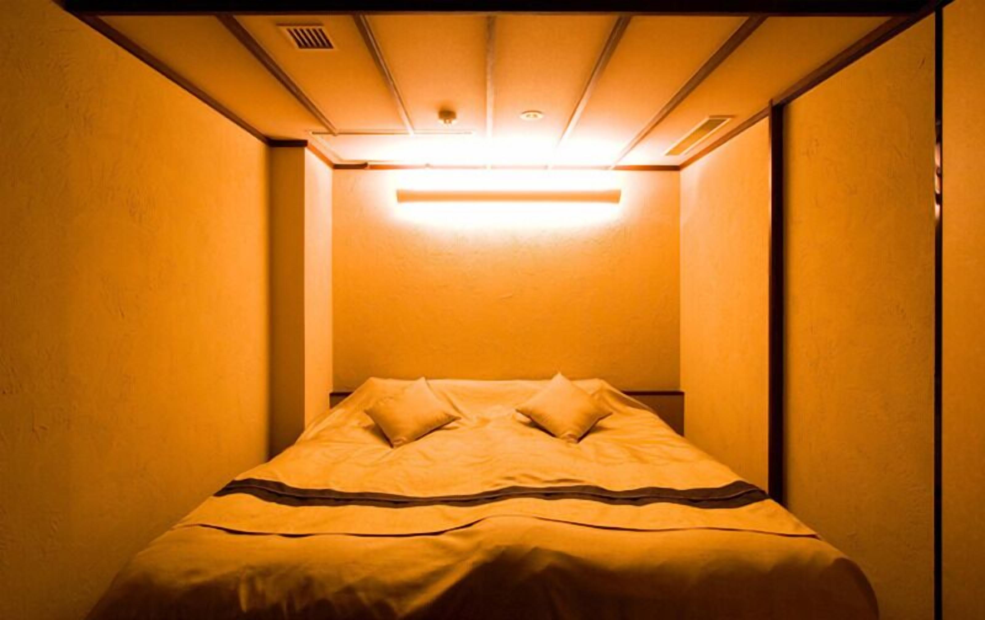 Una habitación de hotel estrecha con una cama individual, cabecera y una lámpara en el techo.