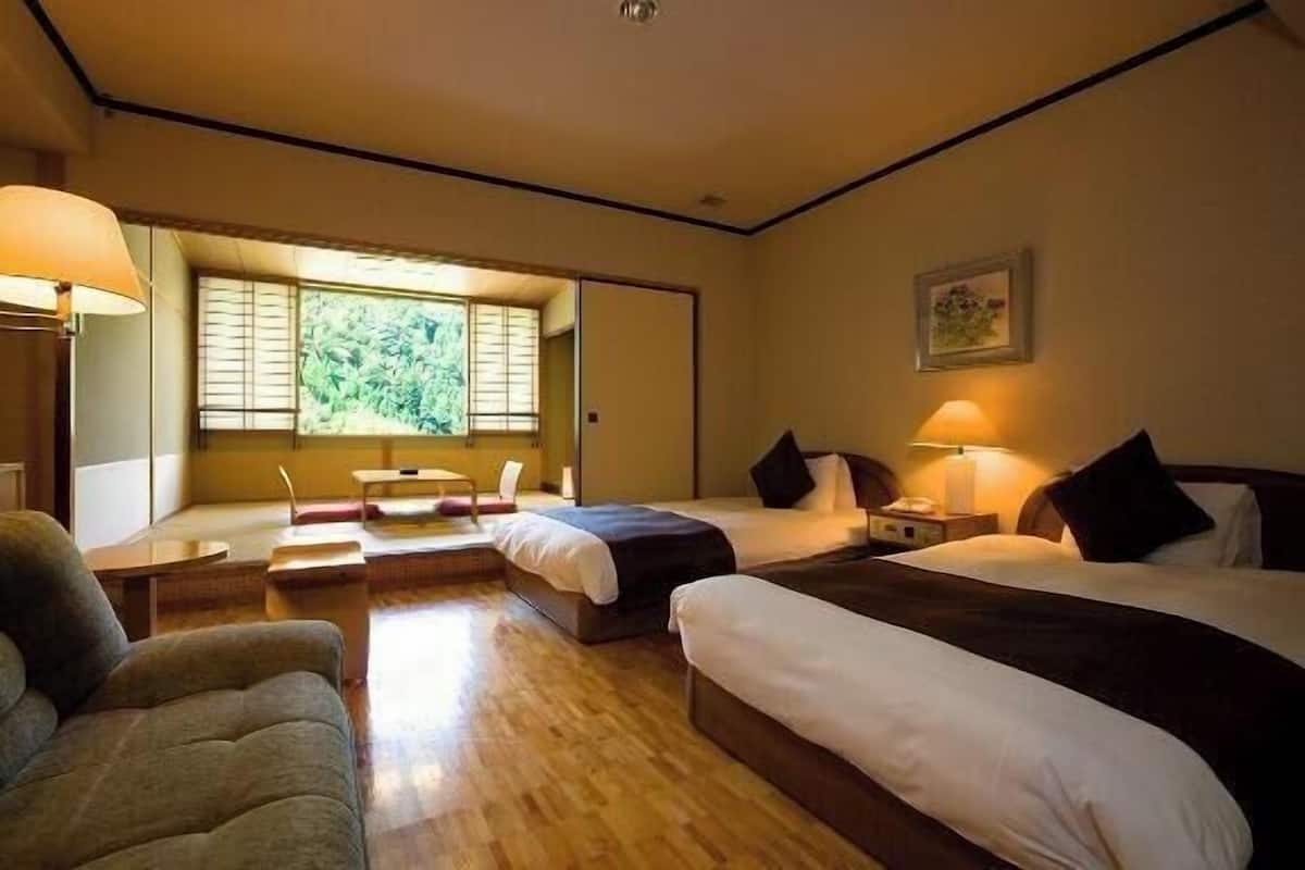 A hotel room with two beds, a sofa, a desk, and a window with a view of greenery.