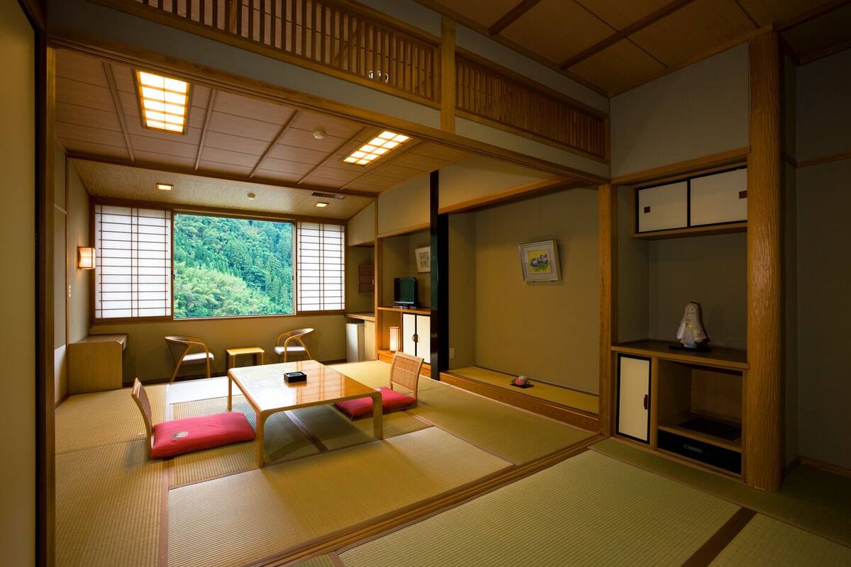 A traditional Japanese room with tatami flooring, a low table with cushions, a TV, and a view of greenery through a large window.