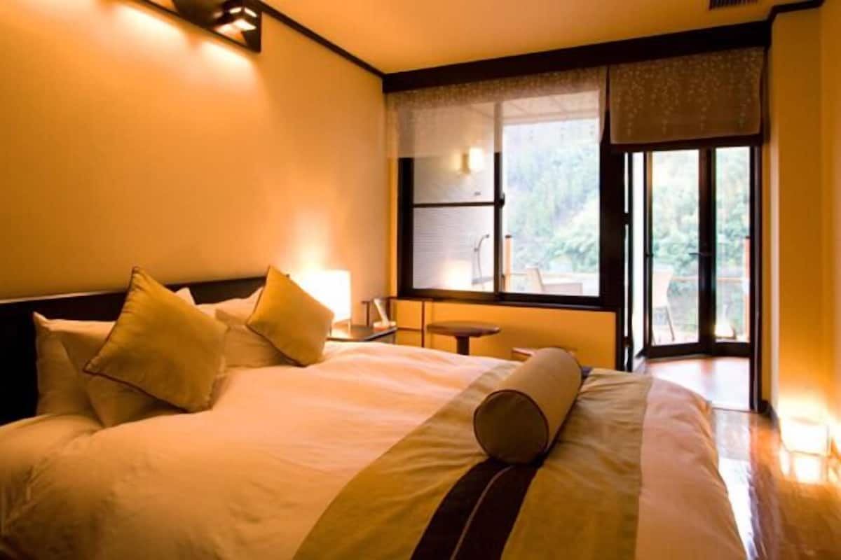 A hotel room with a large bed, a bedside table, a window with a view of greenery, and a small table by the window.