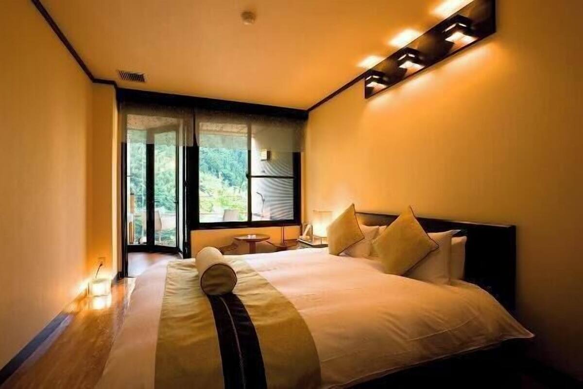 A hotel room with a large bed, a nightstand, a lamp, and a view of greenery through the window.
