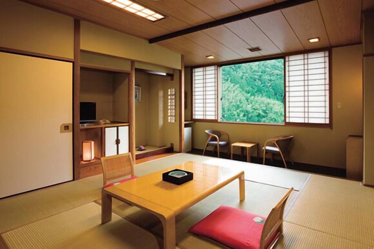 A traditional Japanese room with tatami flooring, a low table with a cushion, a television, and a window with a view of greenery.