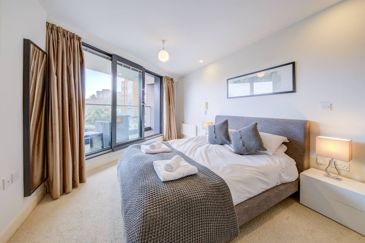 A modern hotel room with a large bed, a bedside table with a lamp, a view of the city through a large window, and a balcony.