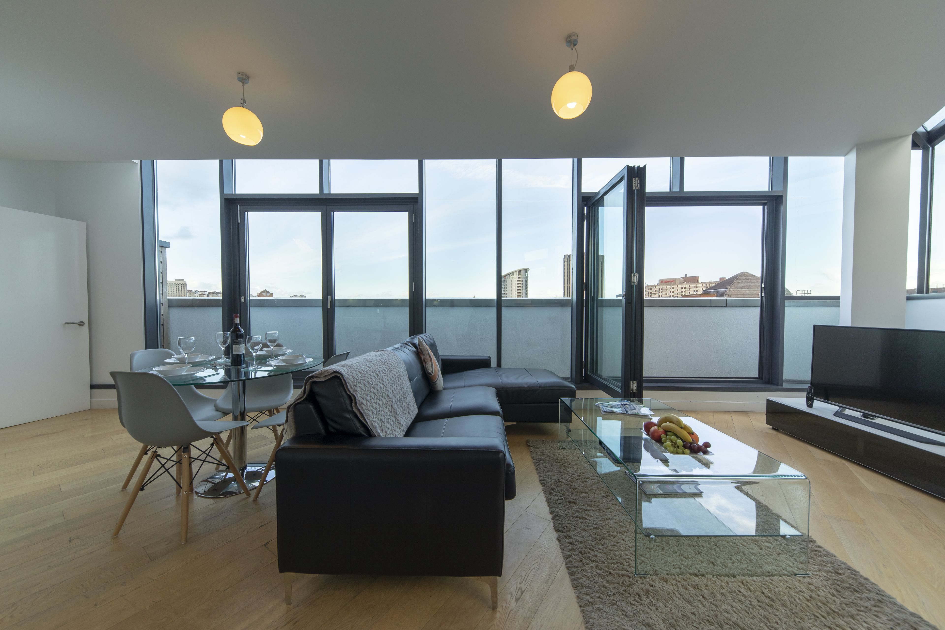 Un salon moderne avec un canapé en cuir noir, une table de salle à manger en verre et un téléviseur à écran plat. De grandes fenêtres offrent une vue sur le paysage urbain.
