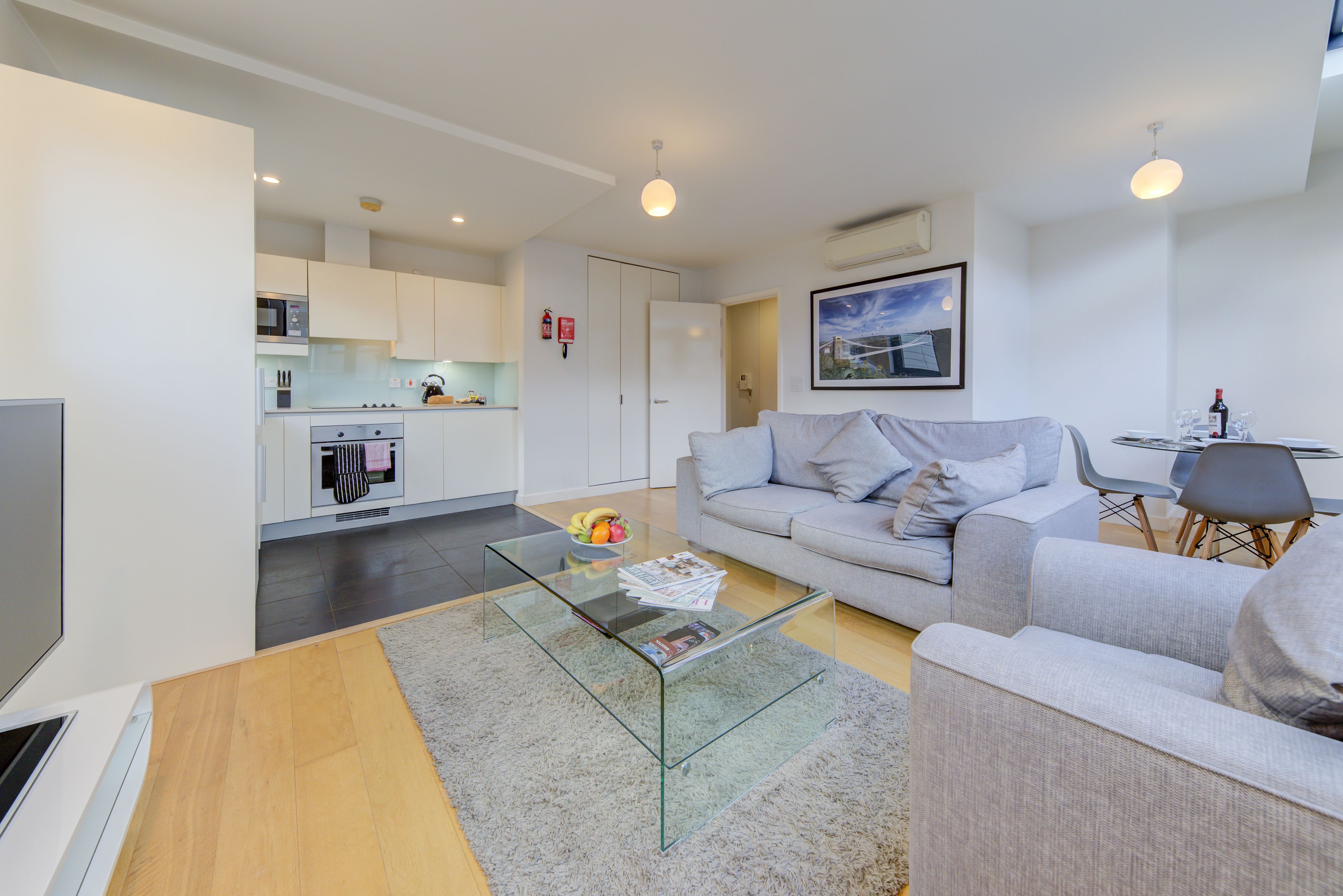 A modern living room with a glass coffee table, grey sofa, and a kitchen in the background.