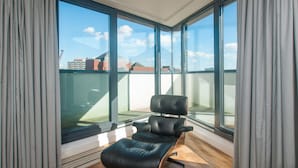 Un balcón moderno con una butaca de cuero, ventanas amplias y vistas a edificios a lo lejos.