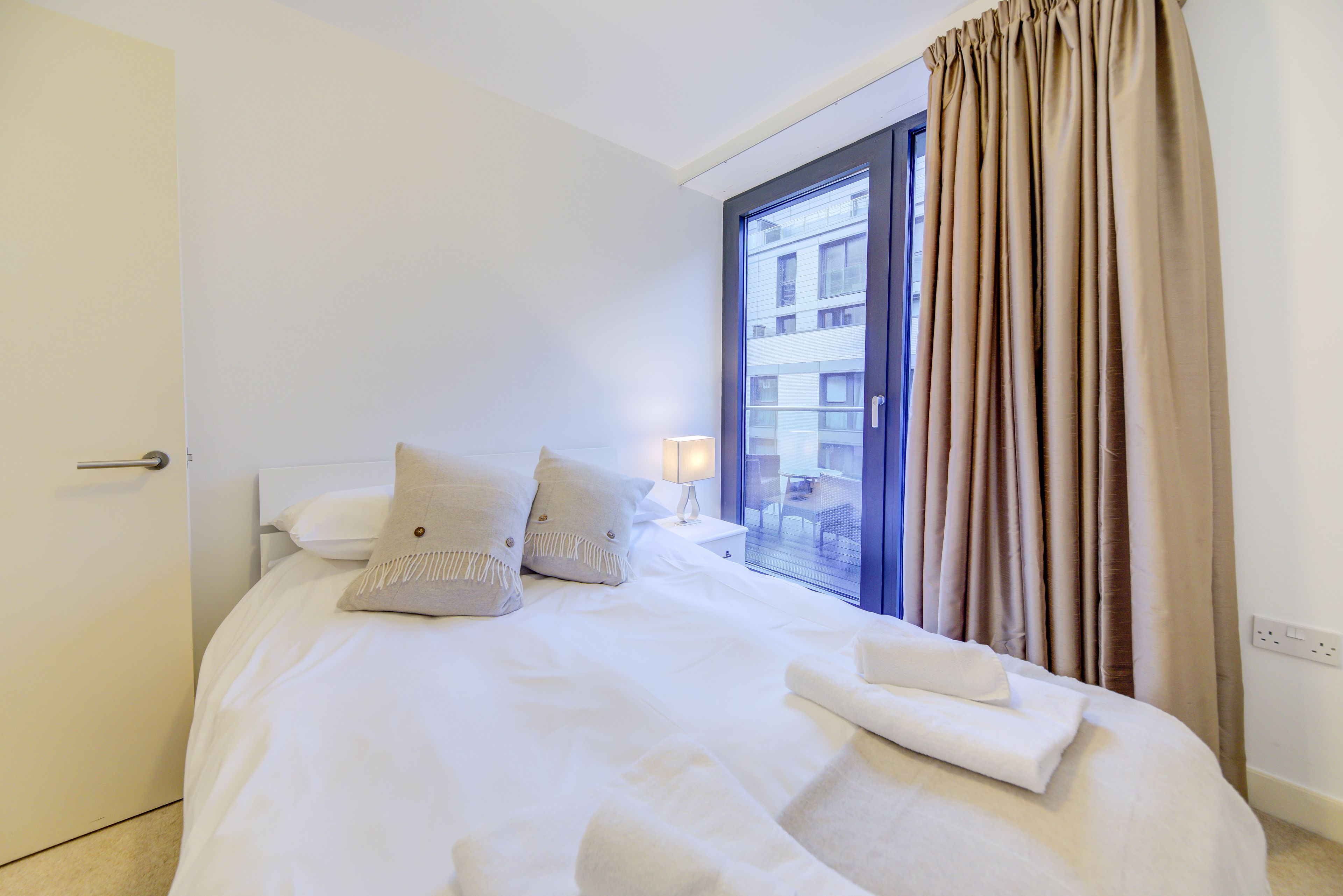 A hotel room with a bed, pillows, a nightstand, a lamp, and a view of the outside through a window with curtains.