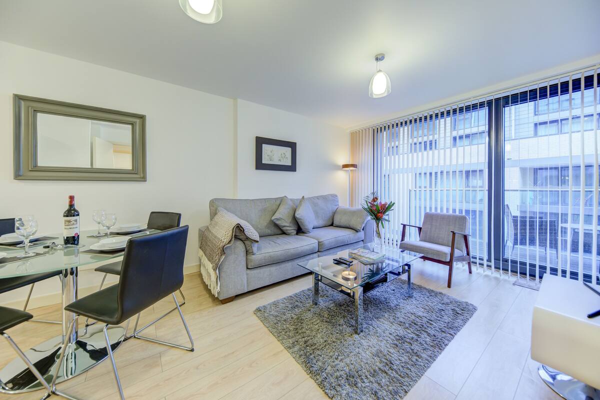 A modern living room with a glass dining table, grey sofa, and a large window with blinds.