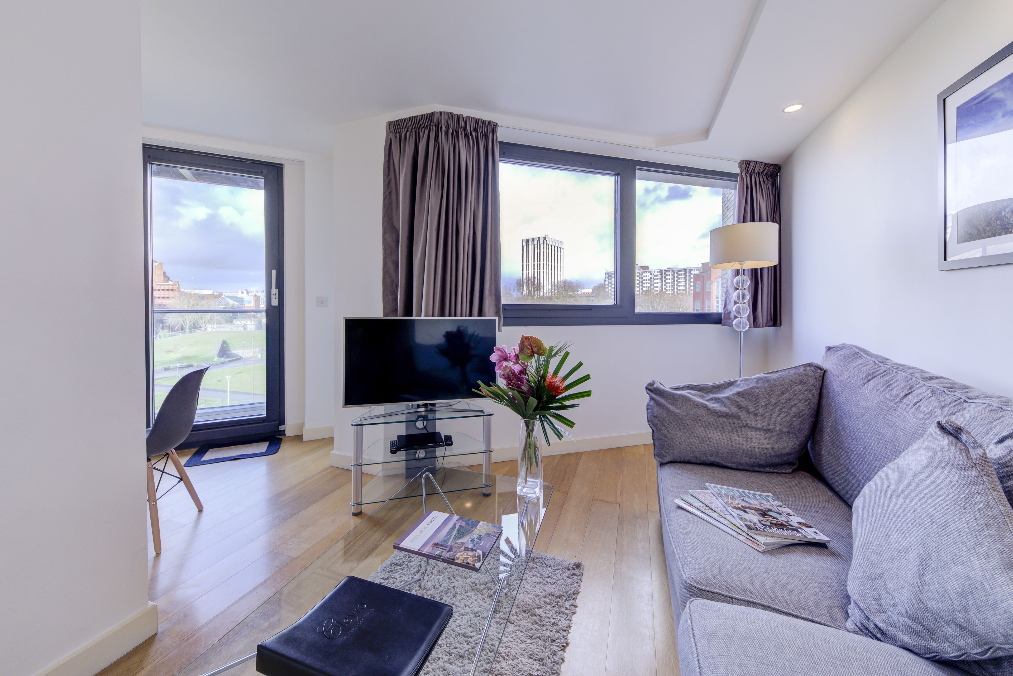 A modern living room with a grey sofa, a flat-screen TV on a glass table, a vase with flowers, and a view of buildings through large windows.