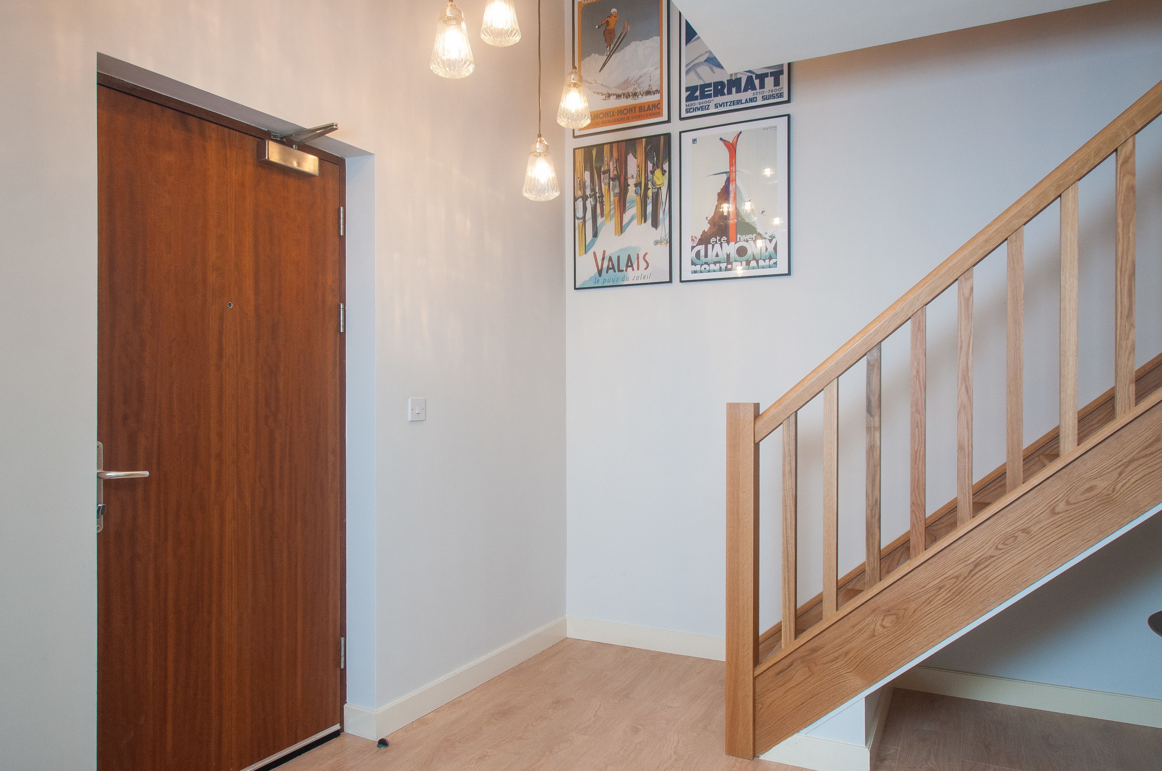 A hallway with a wooden door, a staircase with wooden railings, and four framed posters on the wall.