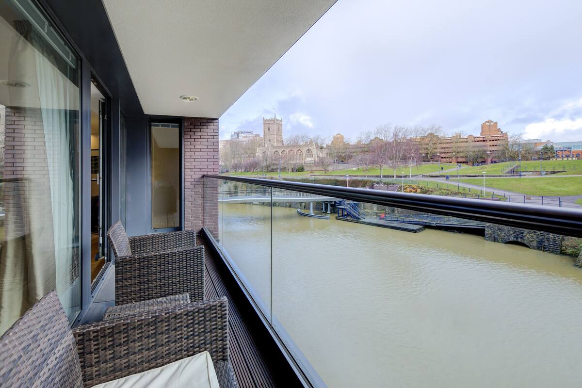 A balcony with wicker furniture overlooking a river, a bridge, and a historic building in the background.