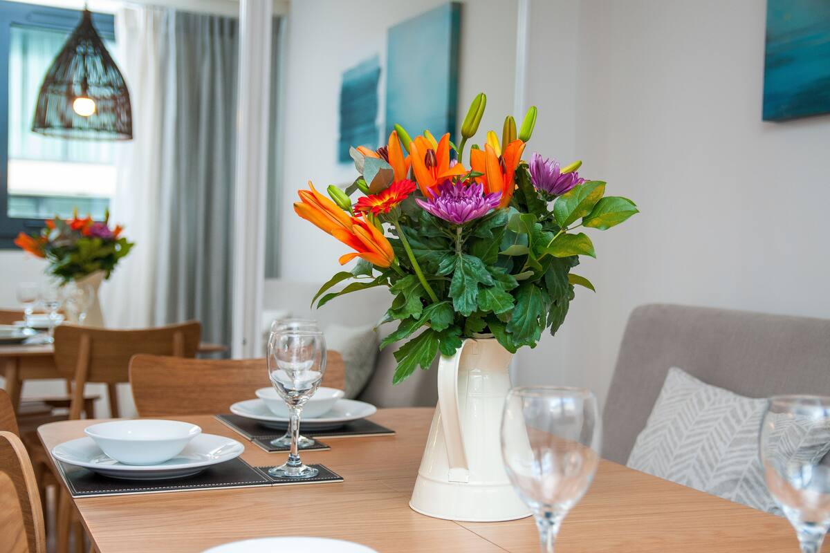 A dining table with a white vase of flowers, a chandelier, and a view of the dining area.