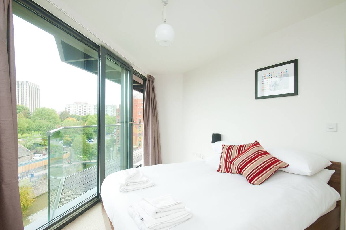 A hotel room with a large window, a bed with white linens, a red and white striped pillow, and a framed picture on the wall.