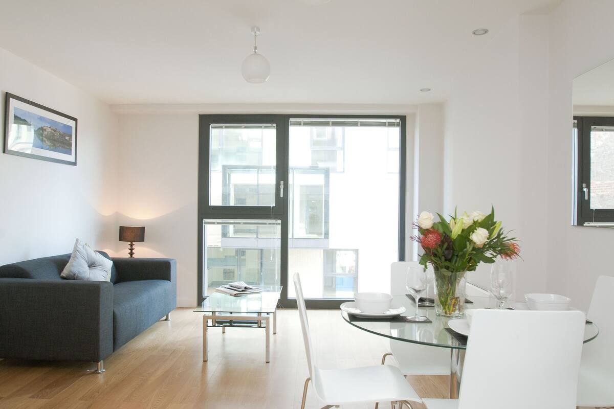 A modern living room with a dark grey sofa, a glass dining table, white chairs, and a vase of flowers.