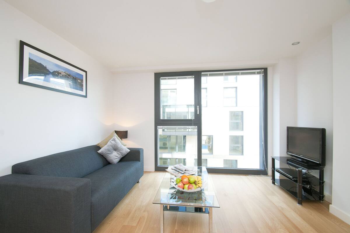 A modern living room with a grey sofa, a glass coffee table with fruit, a flat-screen TV on a stand, and a large window with a city view.
