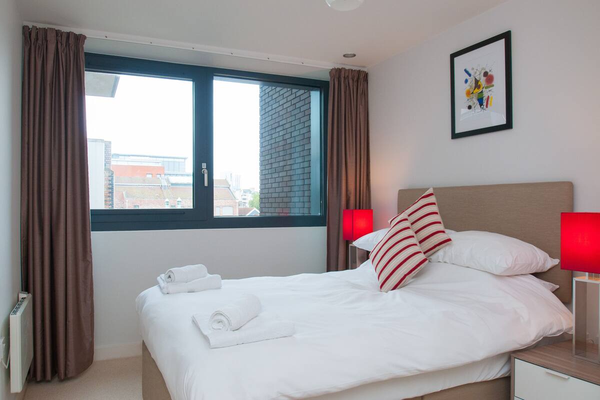 A hotel room with a large bed, a window with curtains, a red lamp, and a framed picture on the wall.