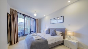 Un dormitorio moderno con una cama grande, una mesita de noche con lámpara y vistas de la ciudad a través de un ventanal amplio.
