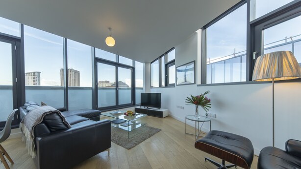 Una sala de estar moderna con un sofá, una mesa de centro de vidrio, un televisor y vistas de la ciudad a través de amplias ventanas.